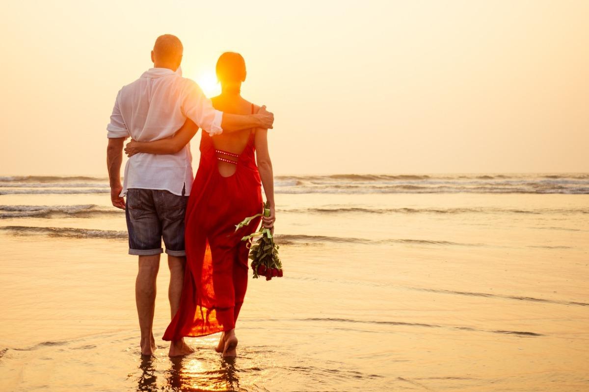 man and woman on beach looking out at the ocean during sunset, woman in a red dress and holding a bouquet of flowers