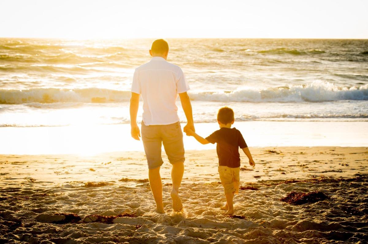 Happy dad and little son walking together on the beach at sunset