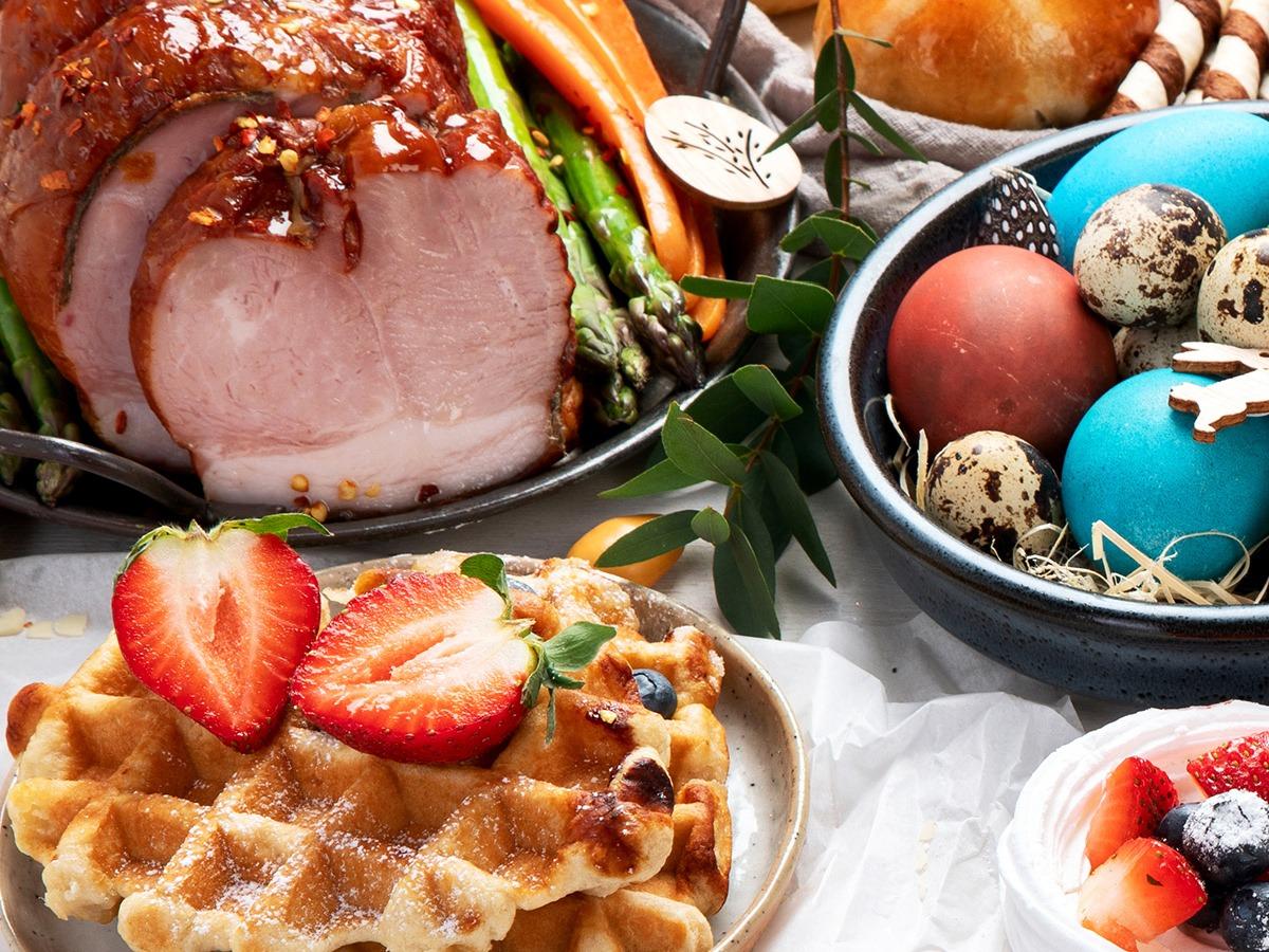 An Easter feast is laid out with a glazed ham taking center stage, sliced to reveal a tender pink interior and garnished with asparagus. A bowl of colorful Easter eggs, some speckled, provides a festive touch. In the foreground, there’s a golden Belgian waffle adorned with fresh strawberries and a dusting of powdered sugar, with blueberries peeking out from the pockets. 