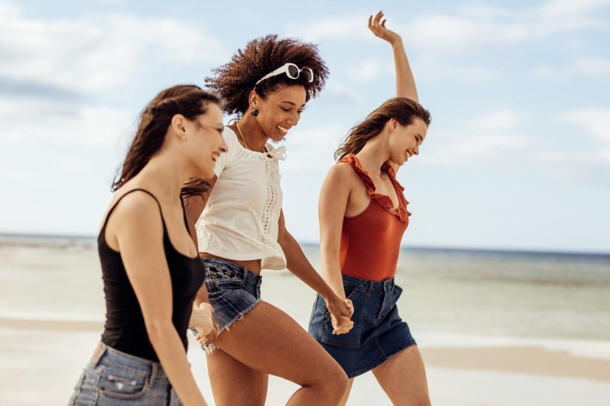 three smiling women laying on a towel on the beach wearing straw hats