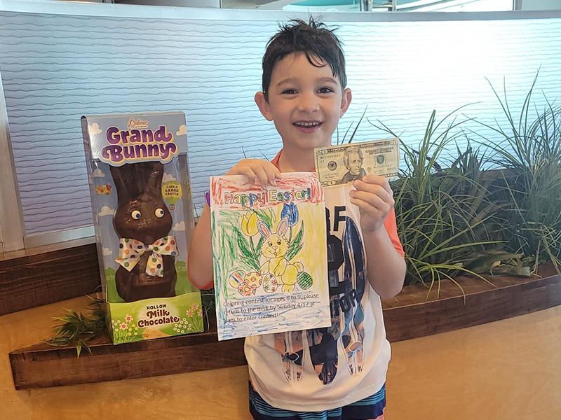 A young boy with wet hair, smiling broadly, is holding a twenty-dollar bill in one hand and a colorful Easter-themed drawing in the other.