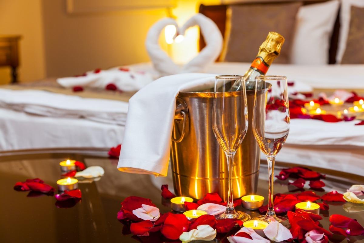 a hotel room with rose petals on bed, towel swans, and a bottle of champagne with two glasses on a table.